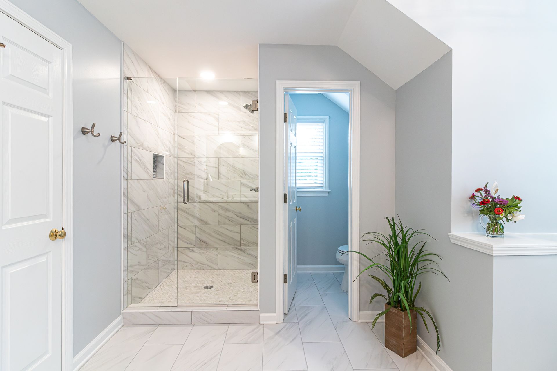 Bright white bathroom with glass shower, tiled walls, potted plant, and open doorway to a blue-lit room