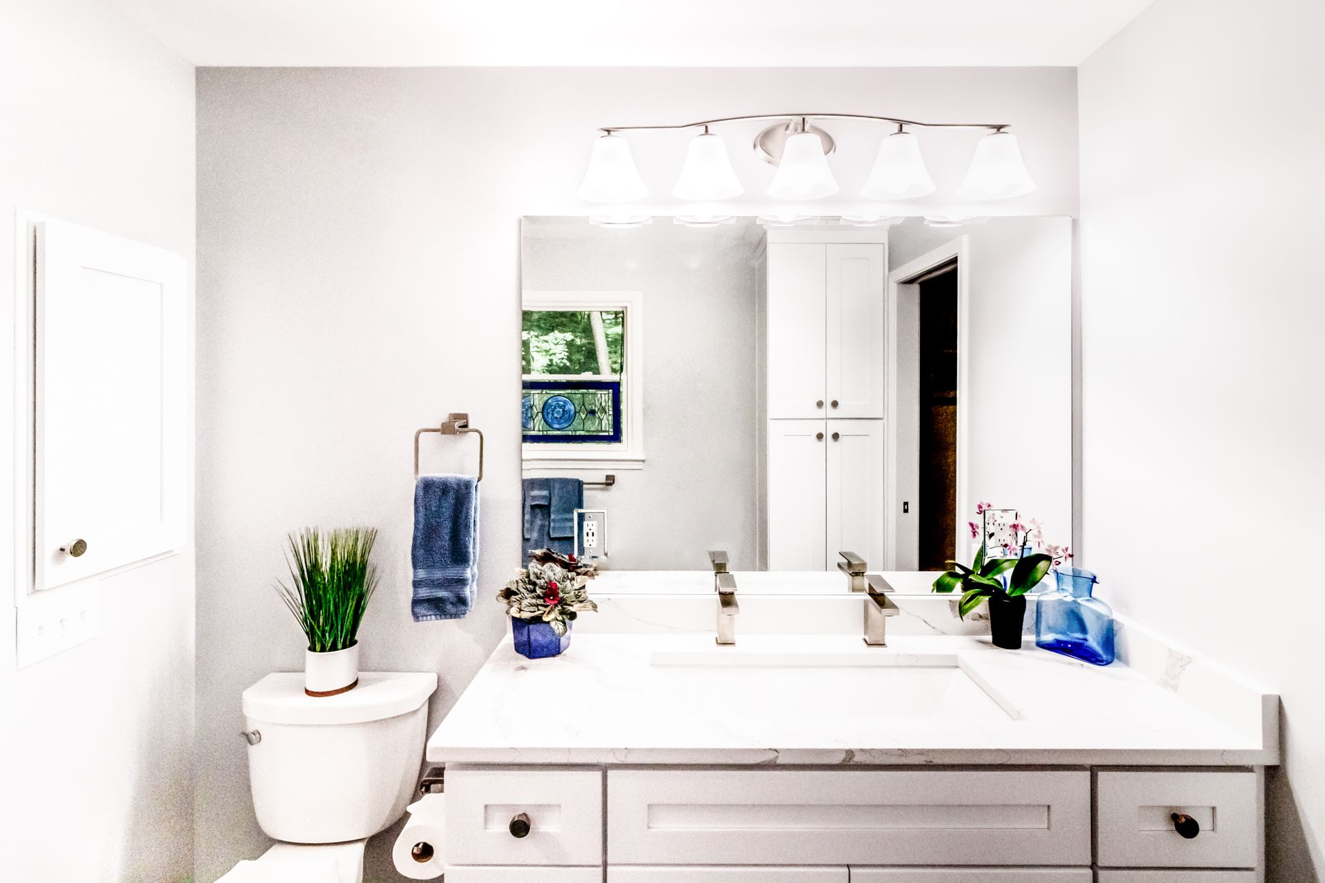 Bright white bathroom with large mirror, vanity, toilet, potted plant, and blue towels