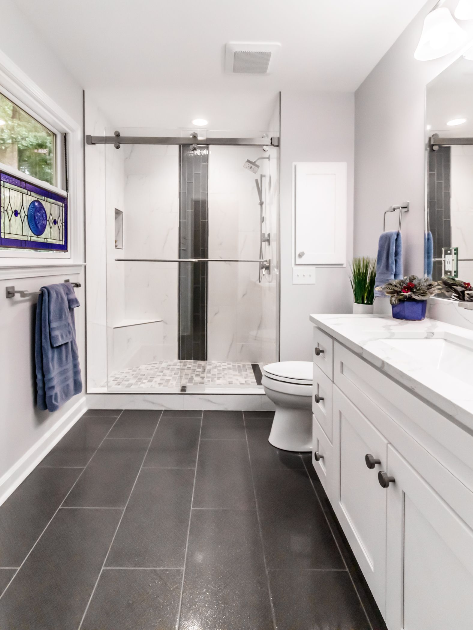 Bright white bathroom with dark tile floor, glass shower, double vanity, and towels by a window.