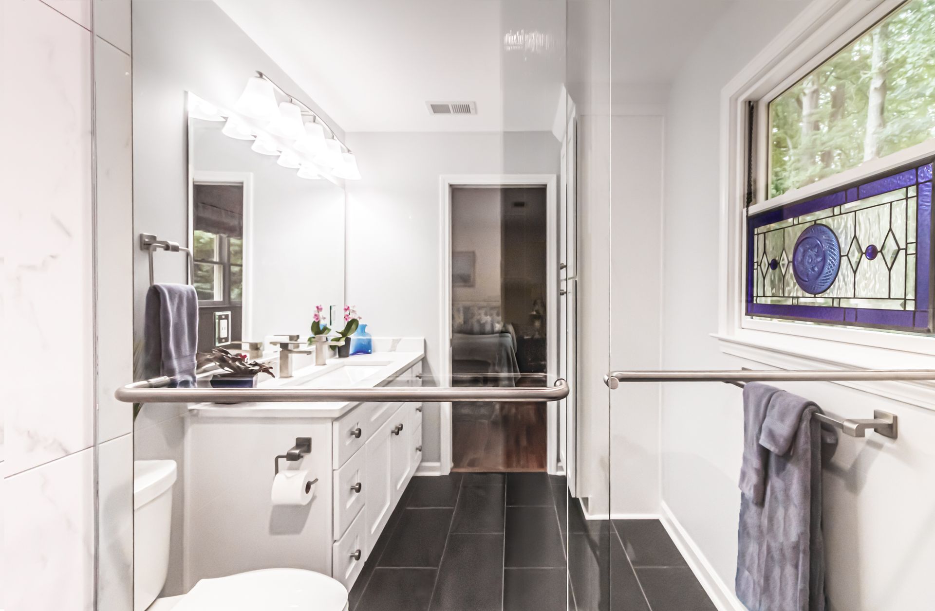 Bright bathroom with white vanity, mirror lights, black tile floor, and a stained-glass window.