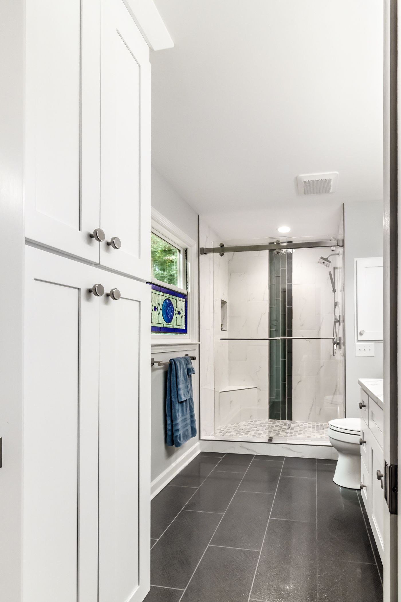 Bright bathroom with white cabinets, dark floor, glass shower, sink, and toilet.