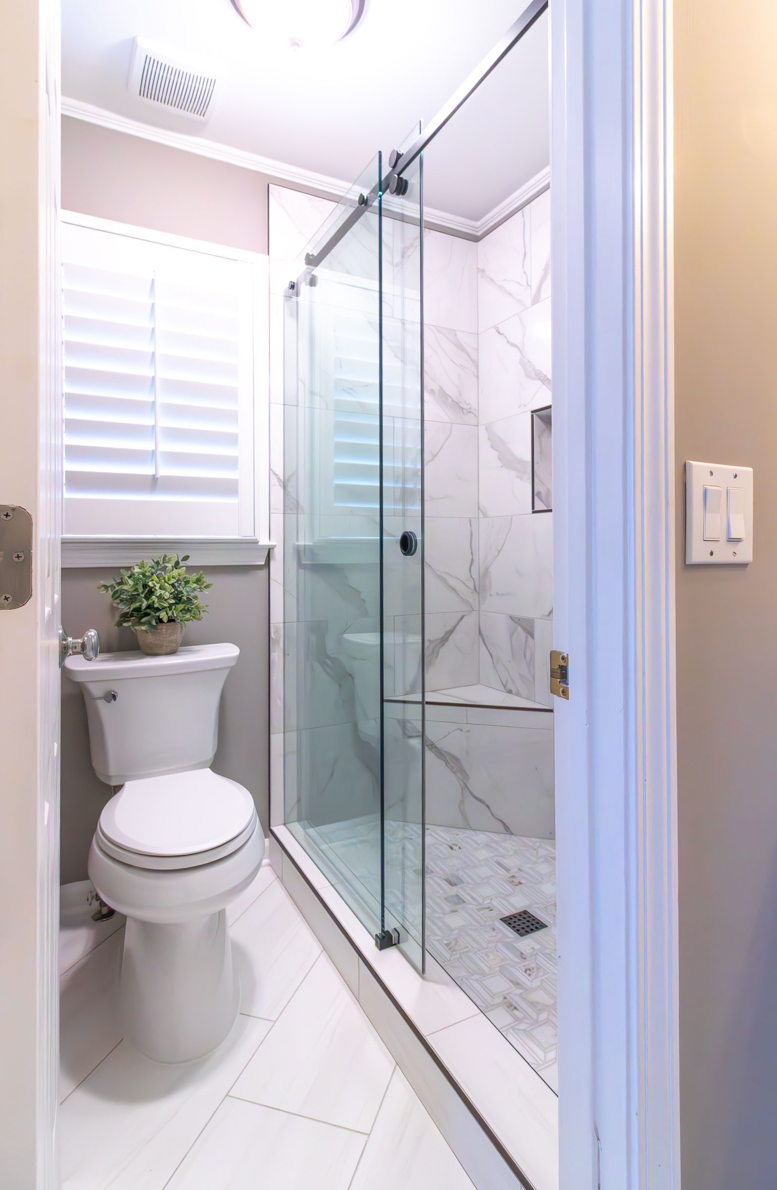 Bright white bathroom with toilet, glass shower enclosure, and window blinds.