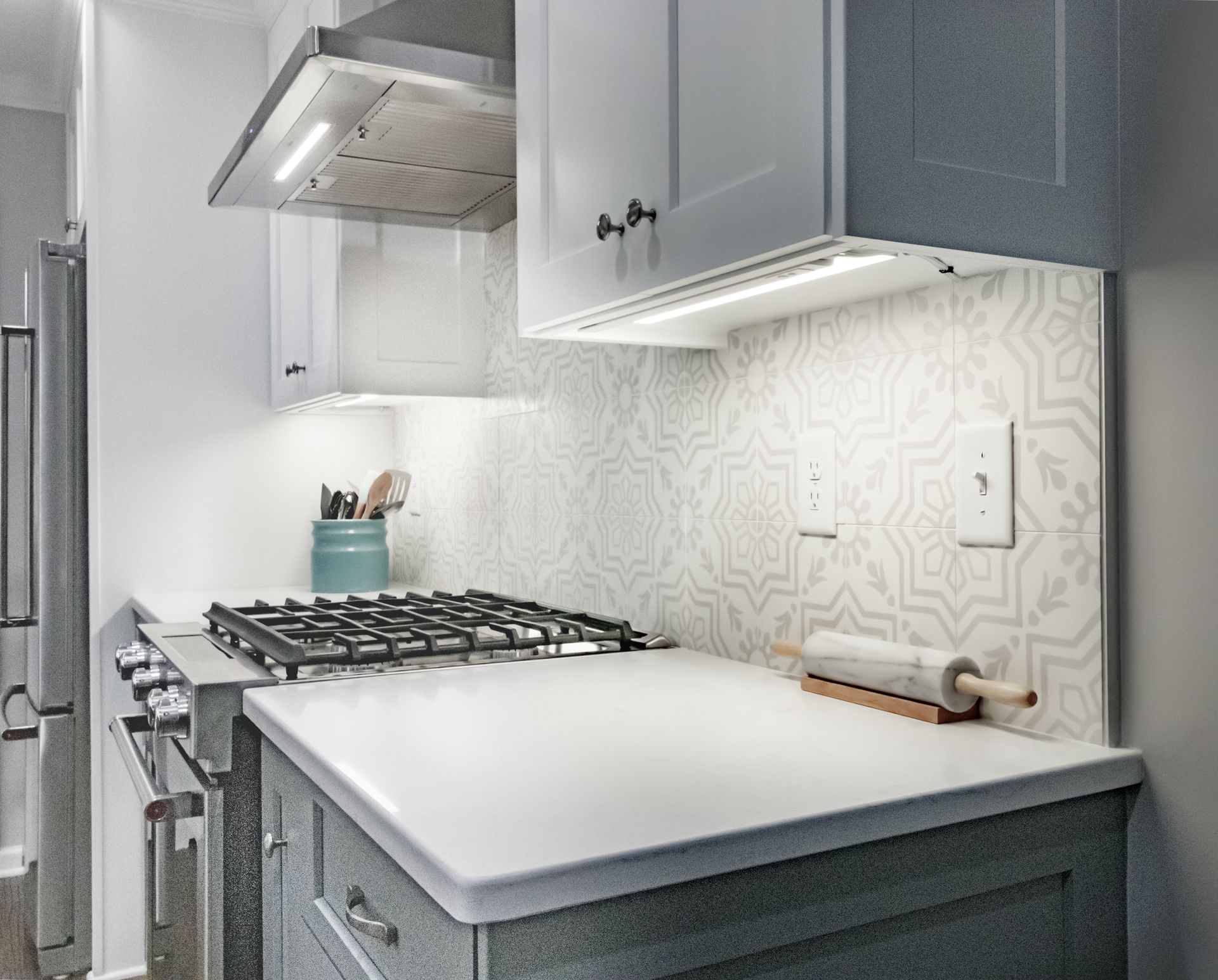 Modern white kitchen counter with gas stove, stainless hood, and blue-gray cabinets, lit by daylight.