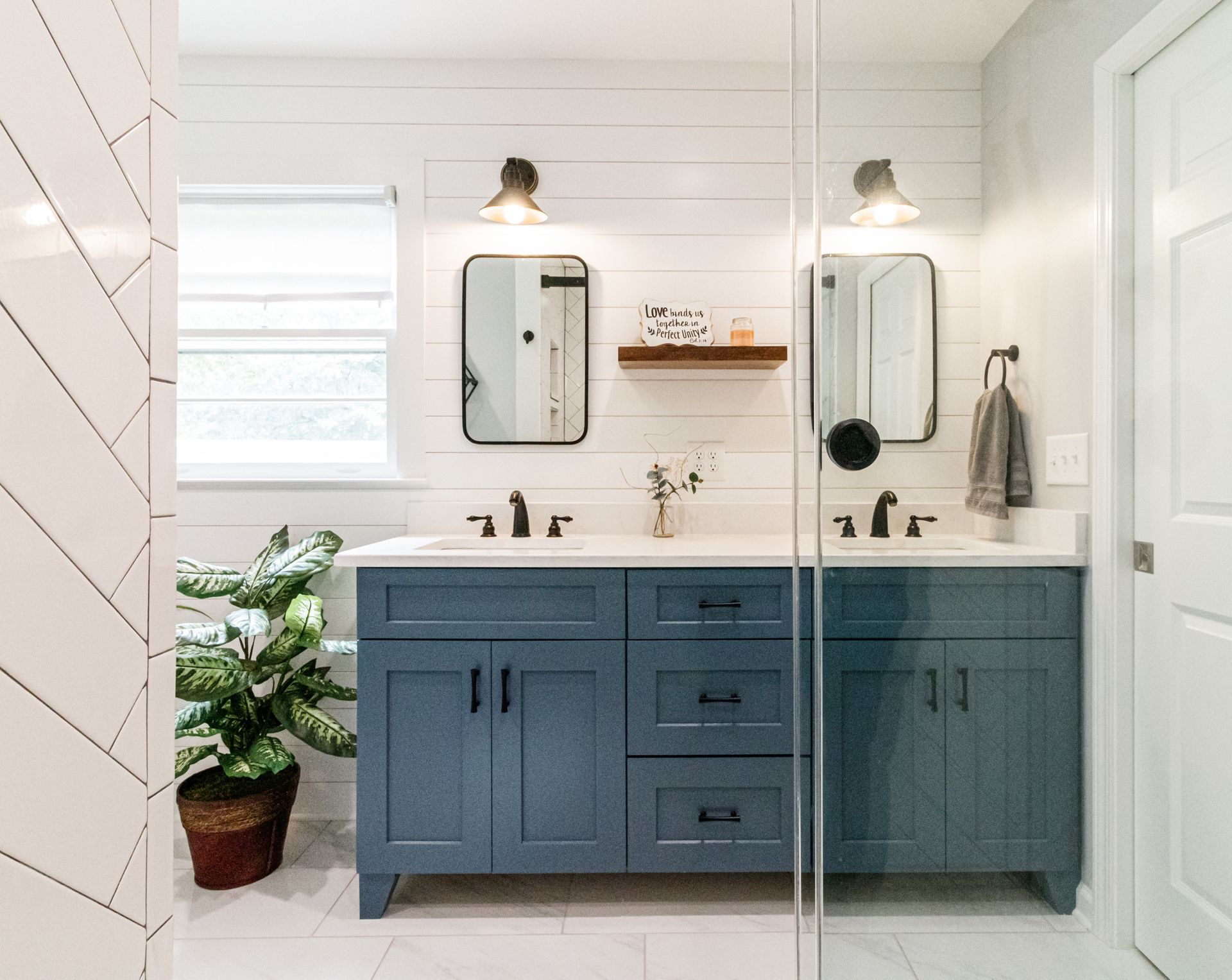 Bright bathroom with blue double vanity, mirrors, sink, and a potted plant by the shower door