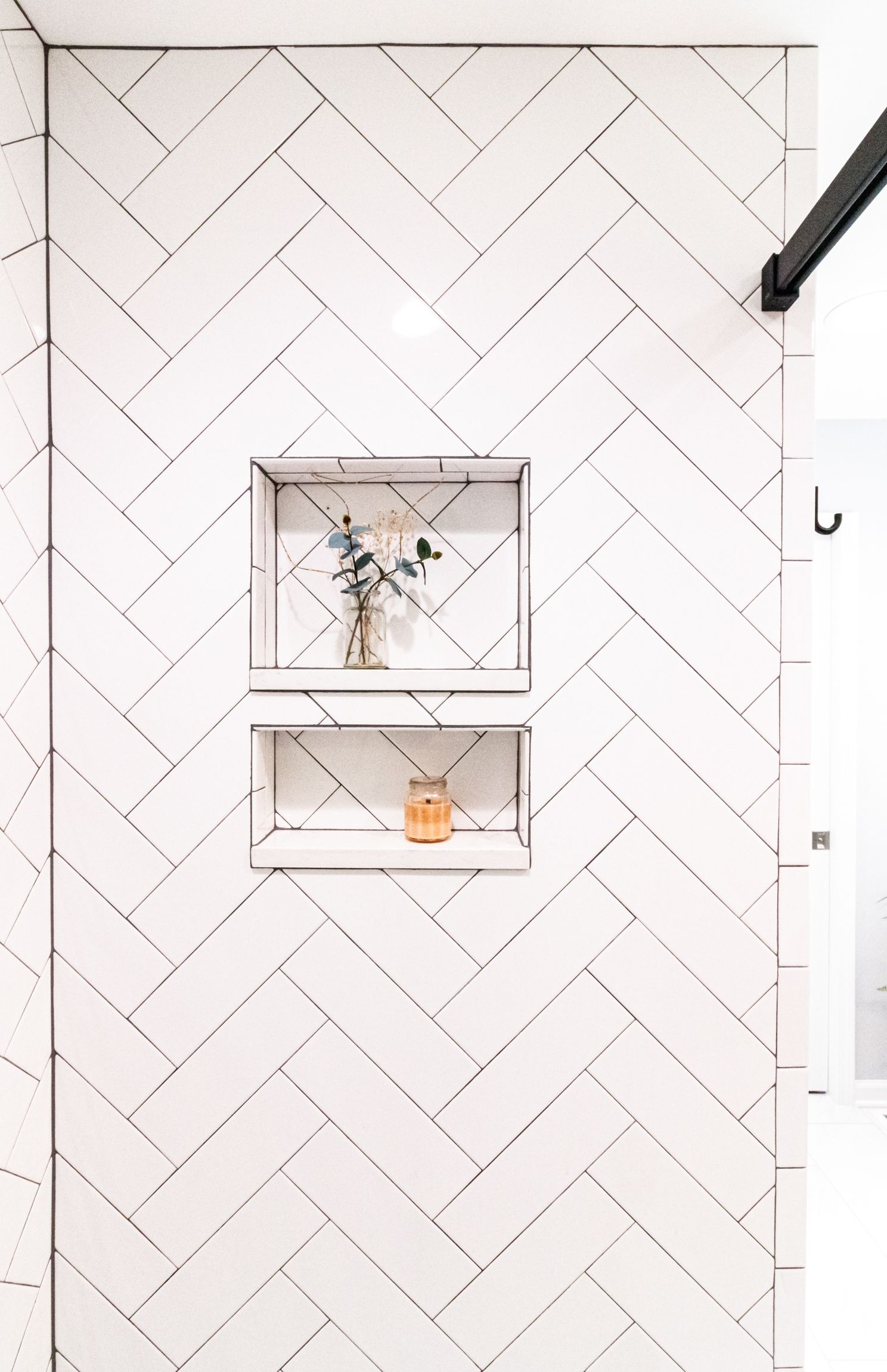 White herringbone-tile wall with two small floating shelves holding a vase and a candle