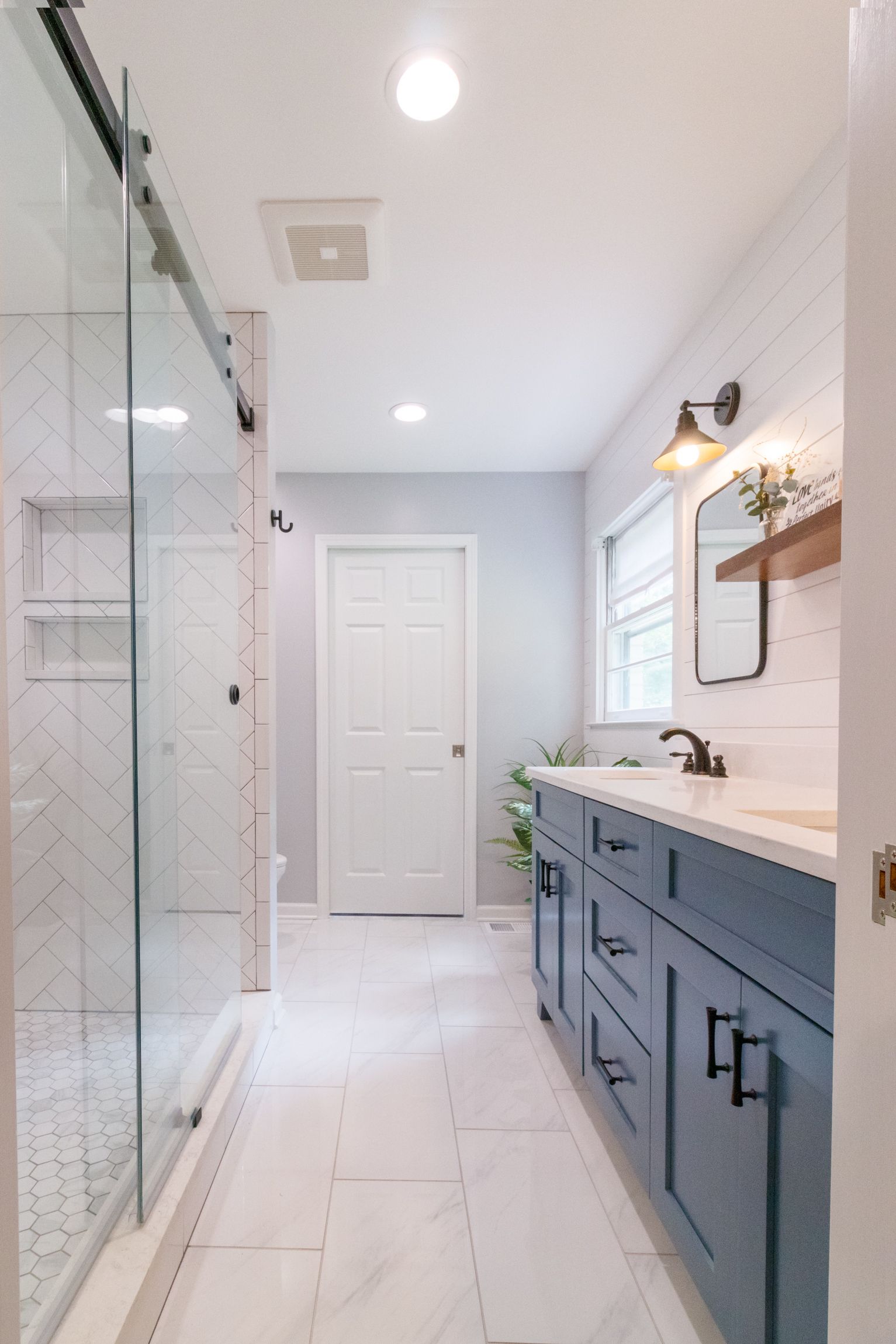Bright bathroom with glass shower, blue vanity, white tile, and a window at the far end.
