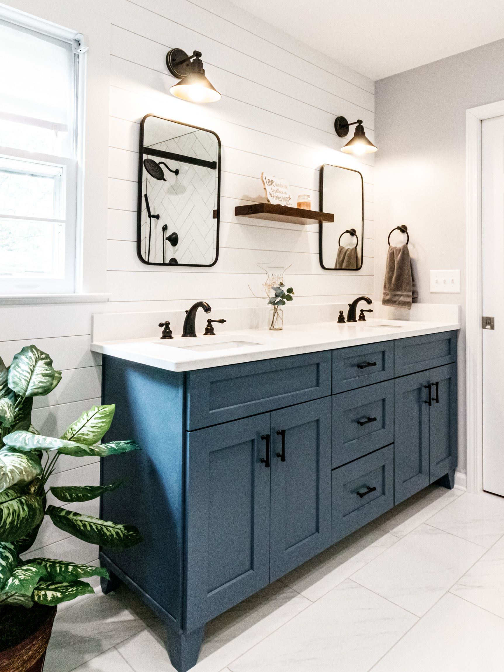 Bright bathroom with blue double vanity, white countertops, mirrors, sconces, and a potted plant near the window