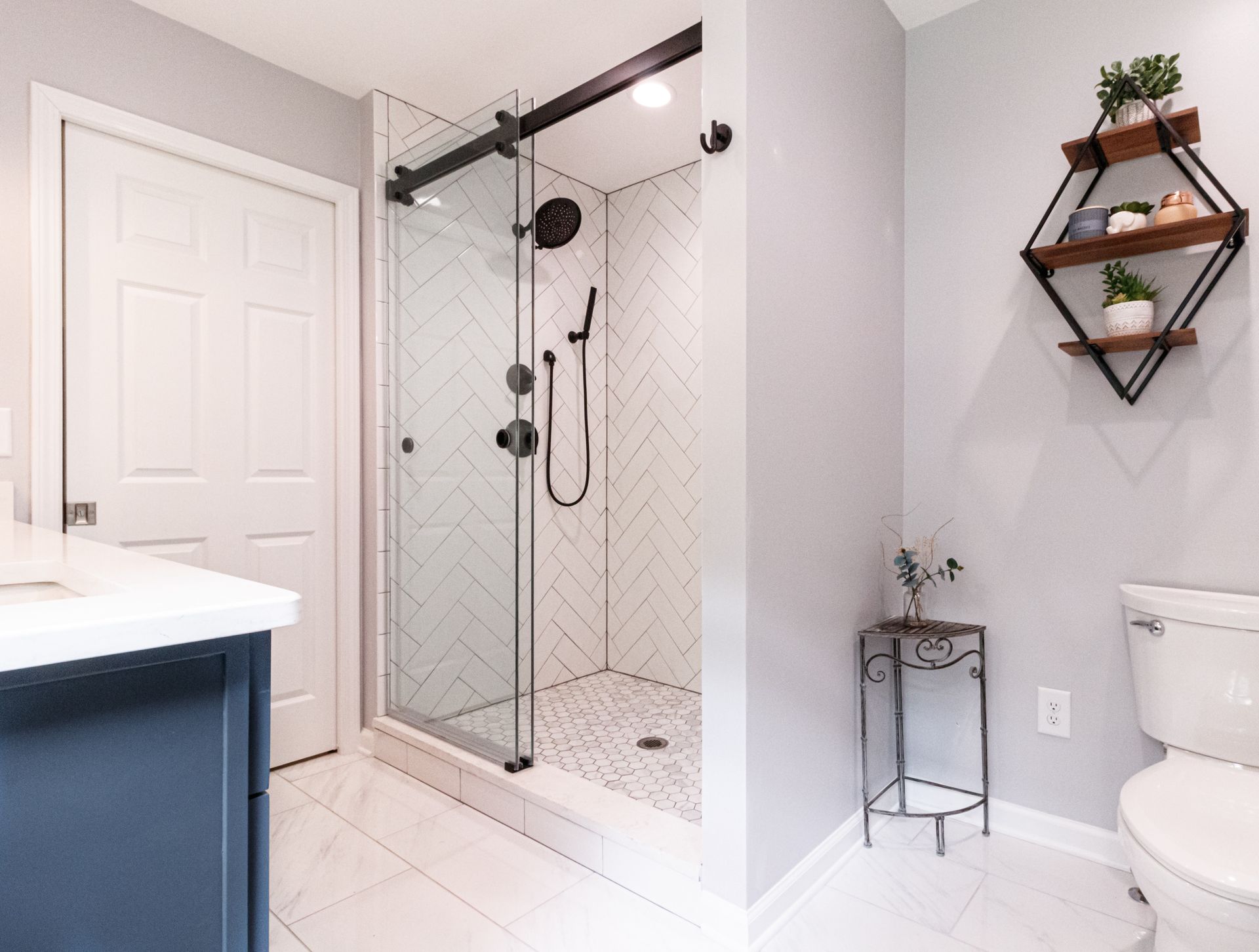Bright bathroom with walk-in glass shower, white tile, vanity edge, and wall shelves with plants