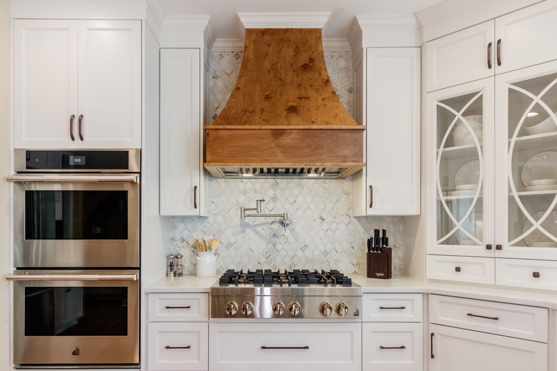 White kitchen with marble backsplash, brass range hood, double ovens, and built-in cabinets