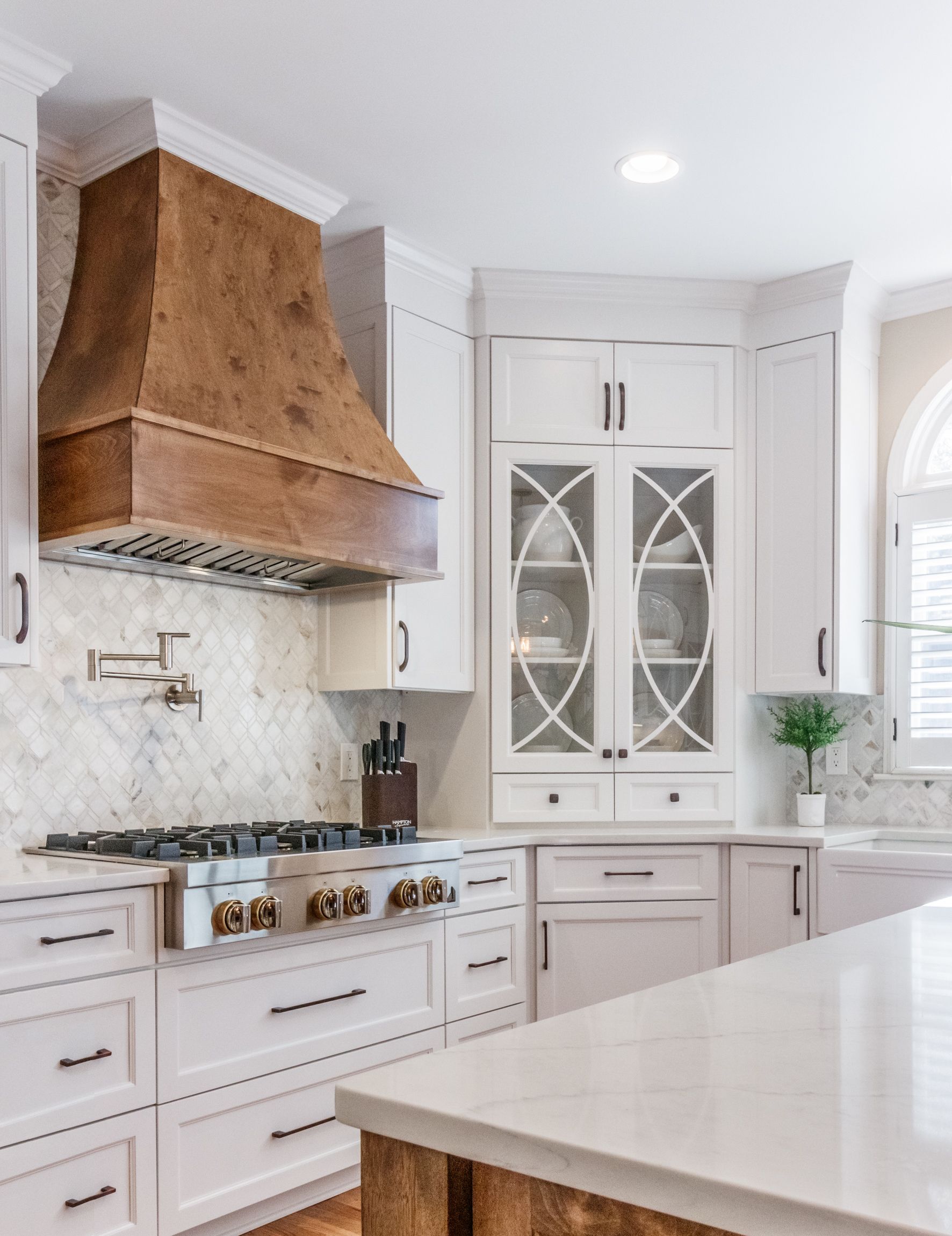 Bright white kitchen with a large range hood, gas stove, and built-in cabinets with glass doors.
