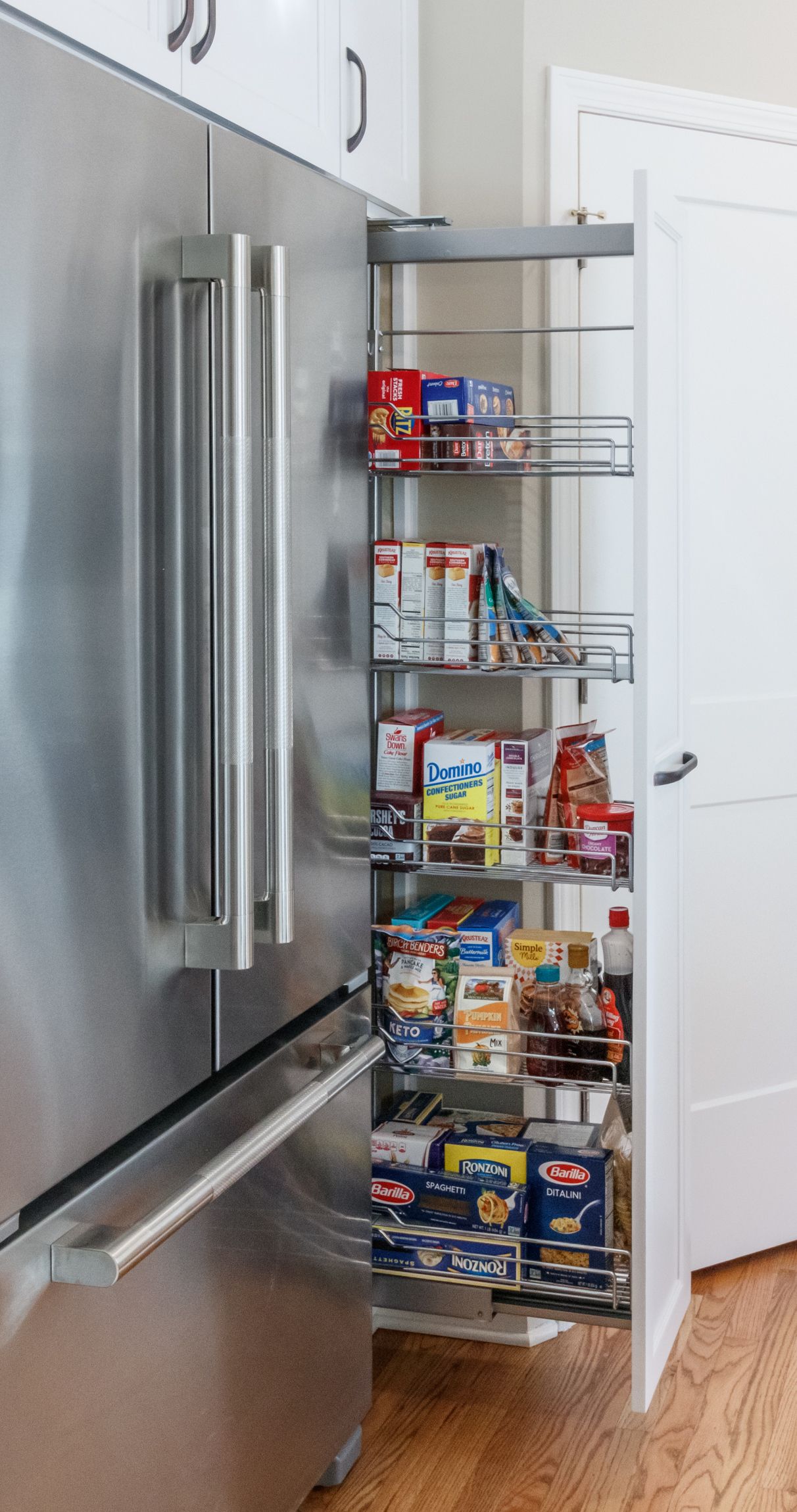 Open pantry door with pull-out wire shelves stocked with snacks and canned goods beside a stainless fridge
