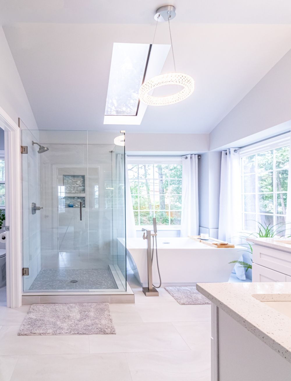 Bright modern bathroom with glass shower, freestanding tub, skylight, and large windows