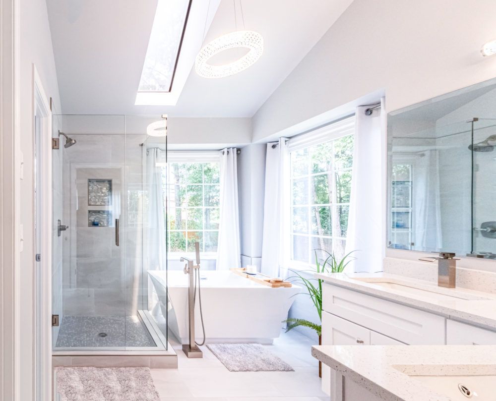 Bright white modern bathroom with glass shower, large windows, freestanding tub, and vanity mirror