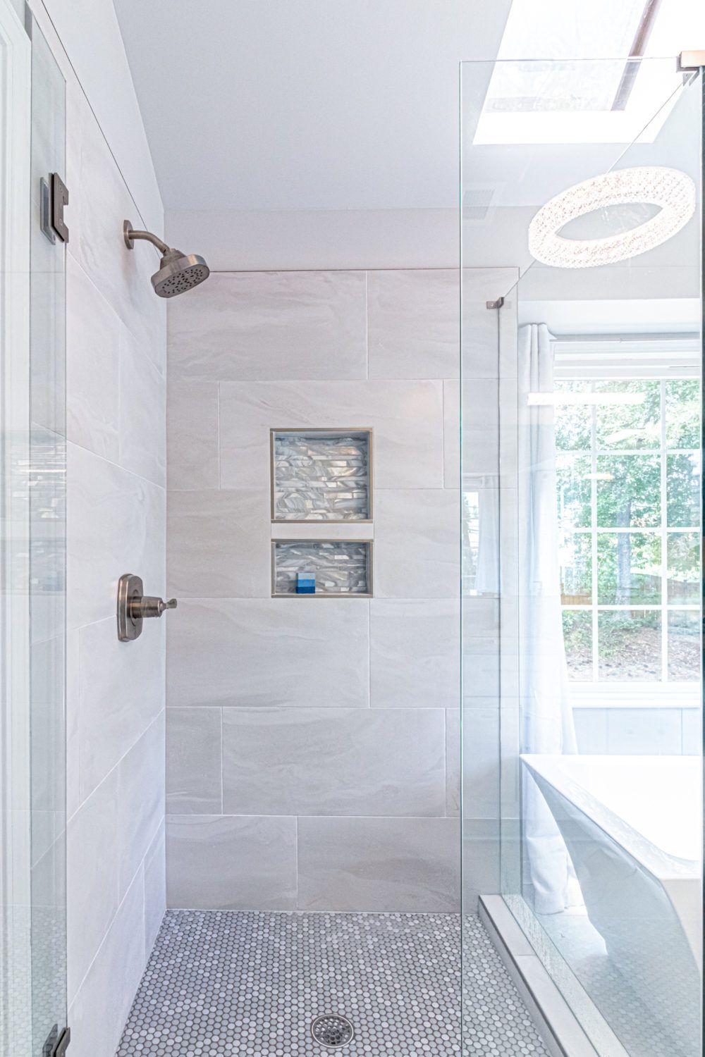 Modern bathroom shower with white tile, pebble floor, built-in niche, and tub by a window
