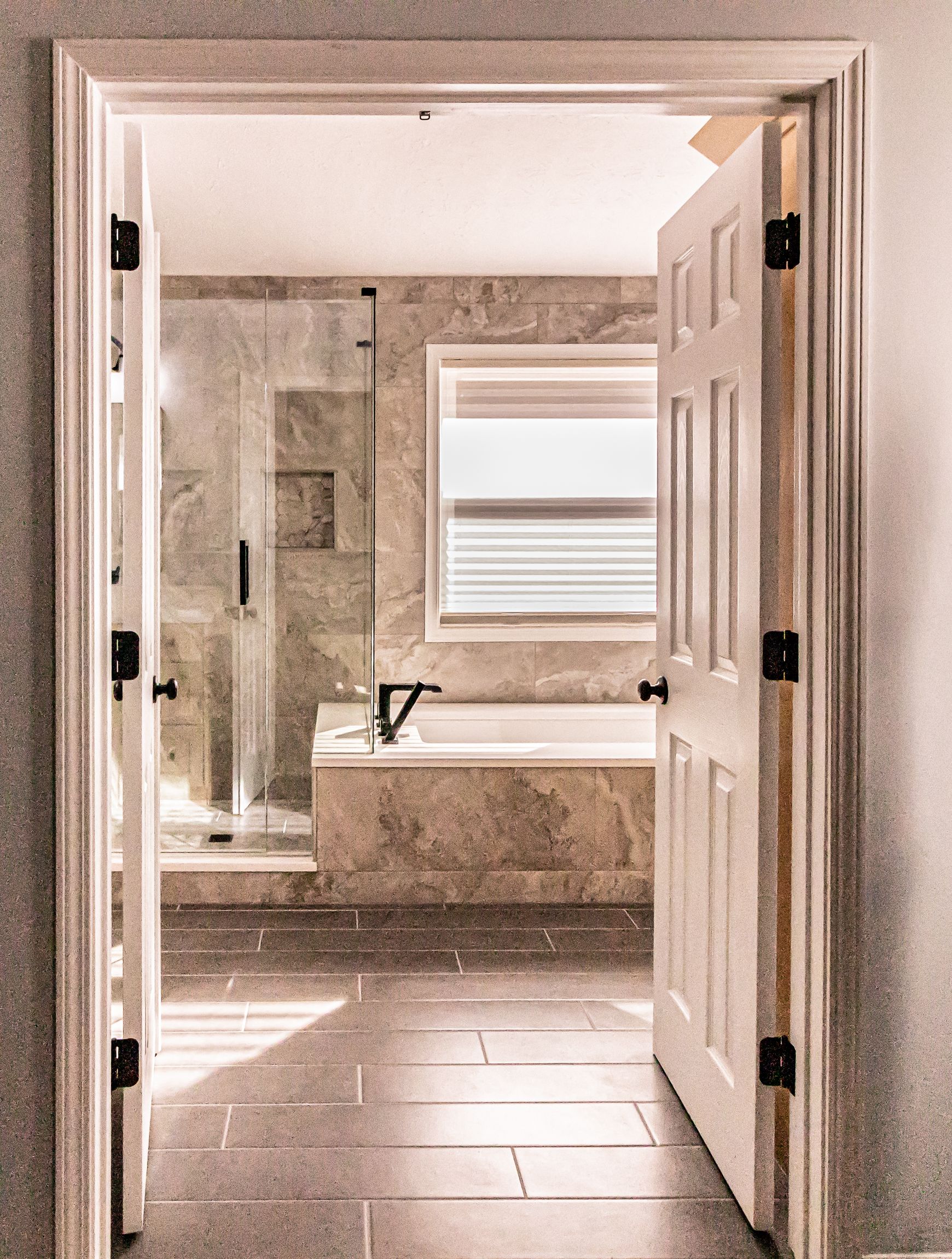 Open bathroom doors revealing a sunlit tub, window, and tiled floor.
