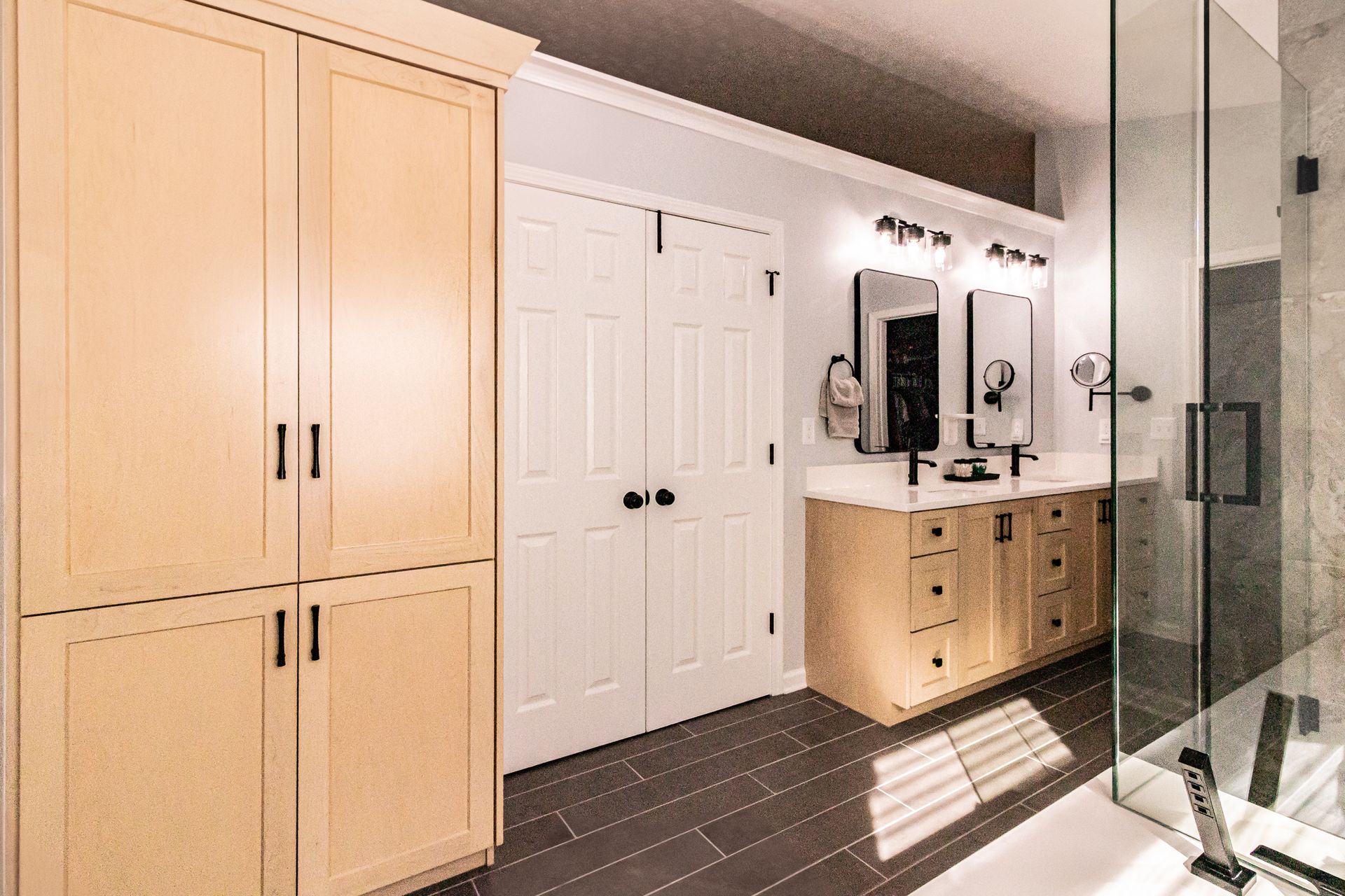 Bright bathroom with light wood cabinets, white double doors, dark tile floor, and glass shower enclosure.