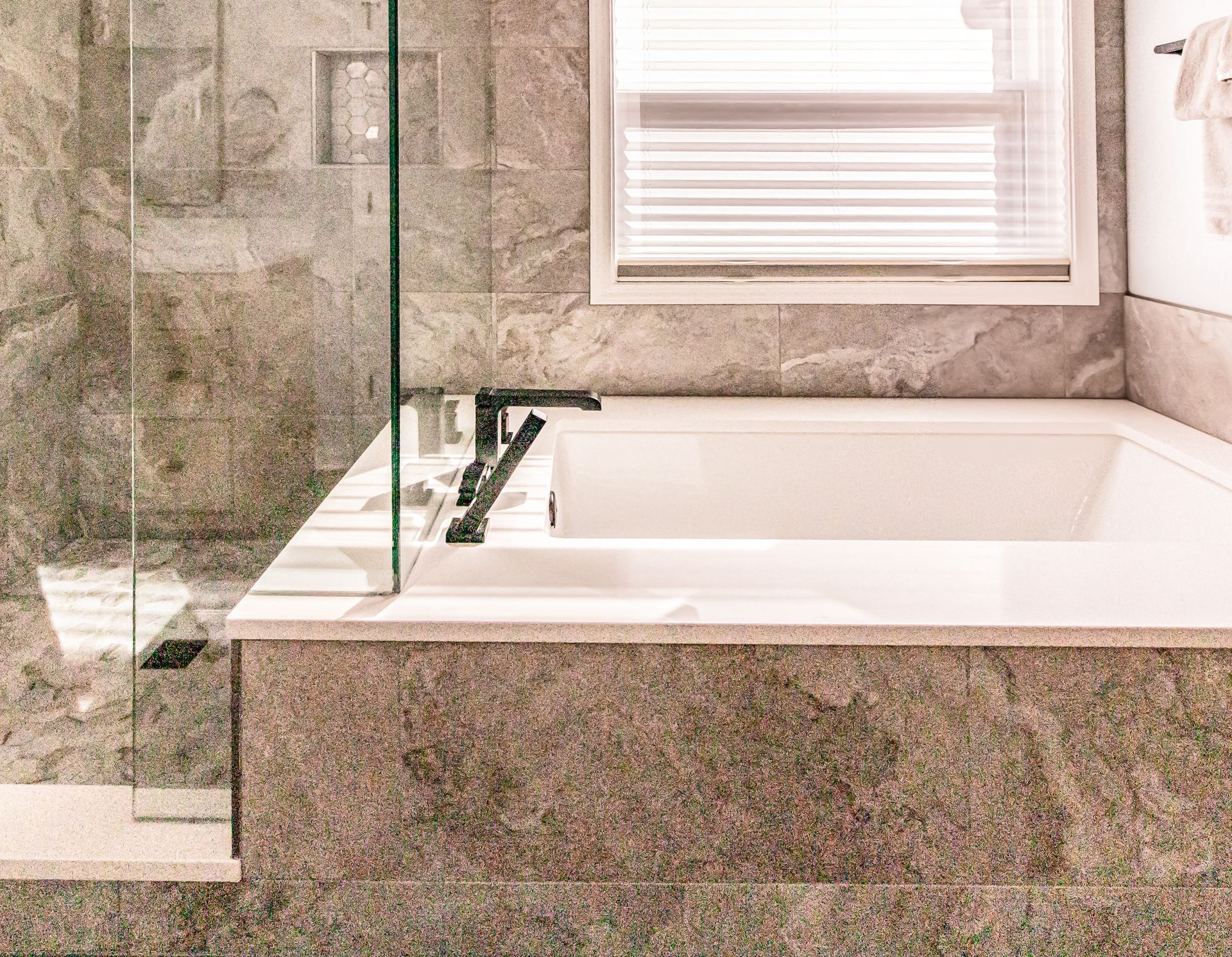 Modern bathroom with a glass-enclosed bathtub, stone tile surround, and a window with blinds.