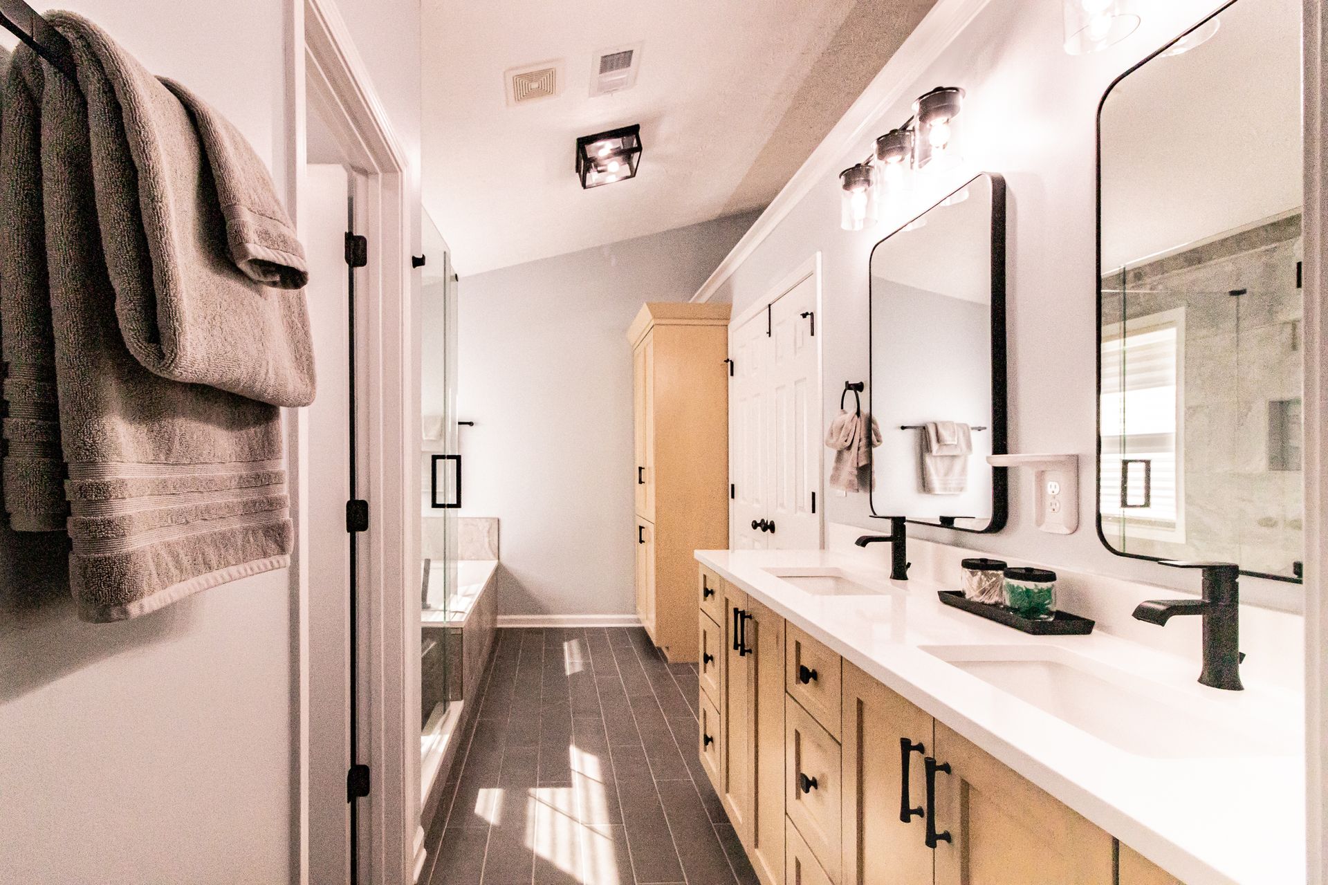 Bright modern bathroom with double vanity, mirrors, towels, and shower visible down the hall