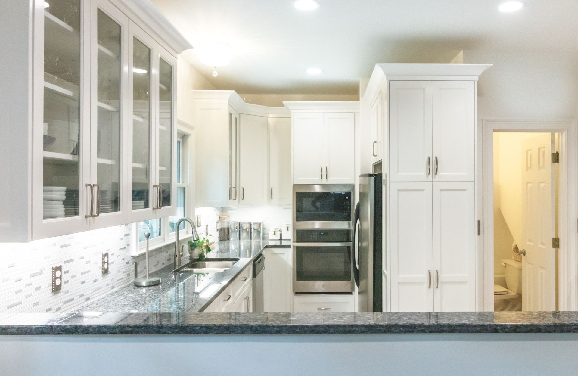 Bright modern white kitchen with glass-front cabinets, granite counters, and built-in ovens