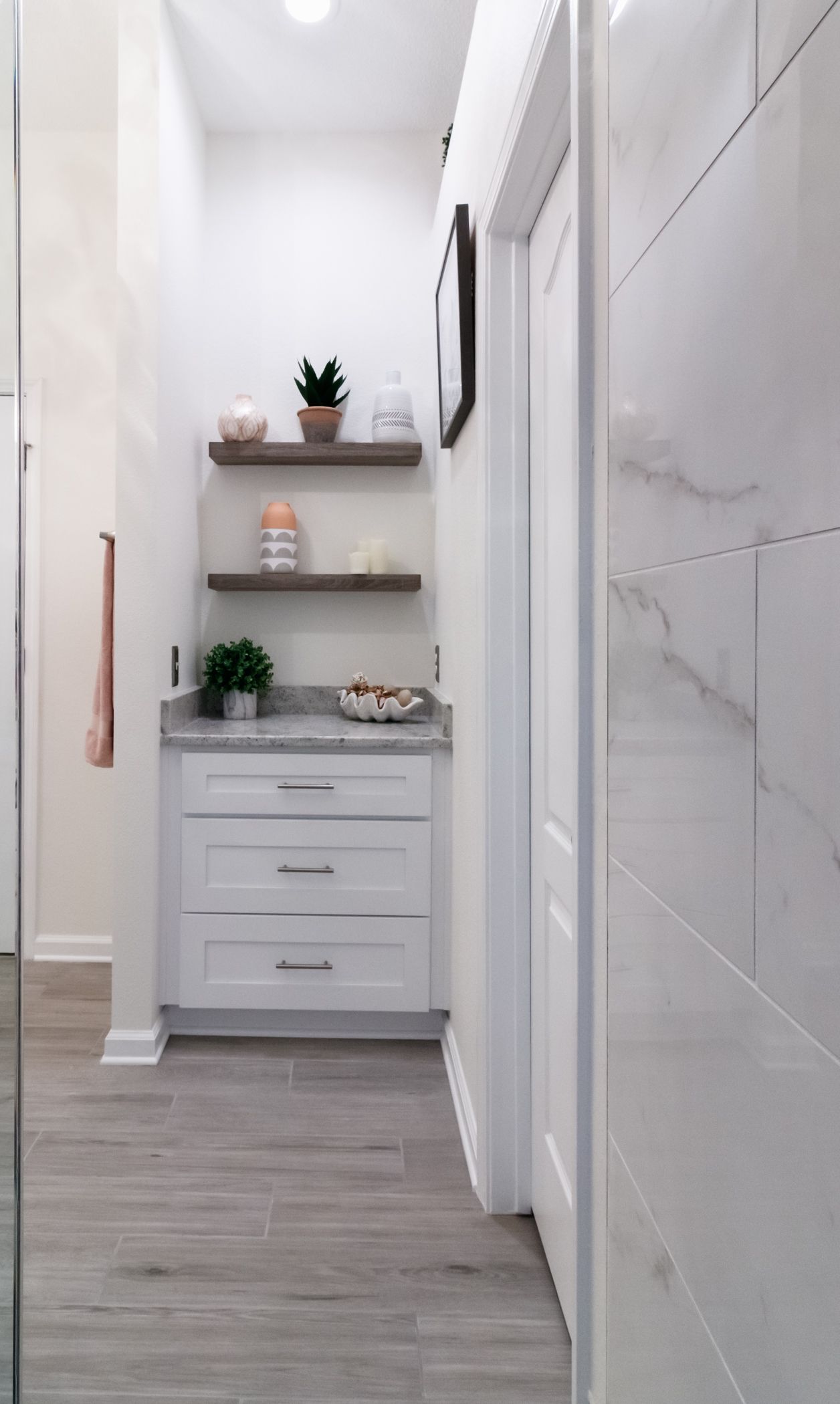 Narrow bathroom hallway with white walls, floating shelves, and a small white cabinet with plants and decor.