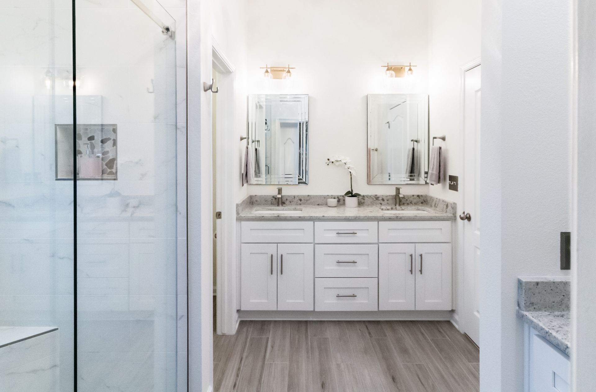 Bright white bathroom with double vanity, mirrors, and glass shower door, viewed through open doorway