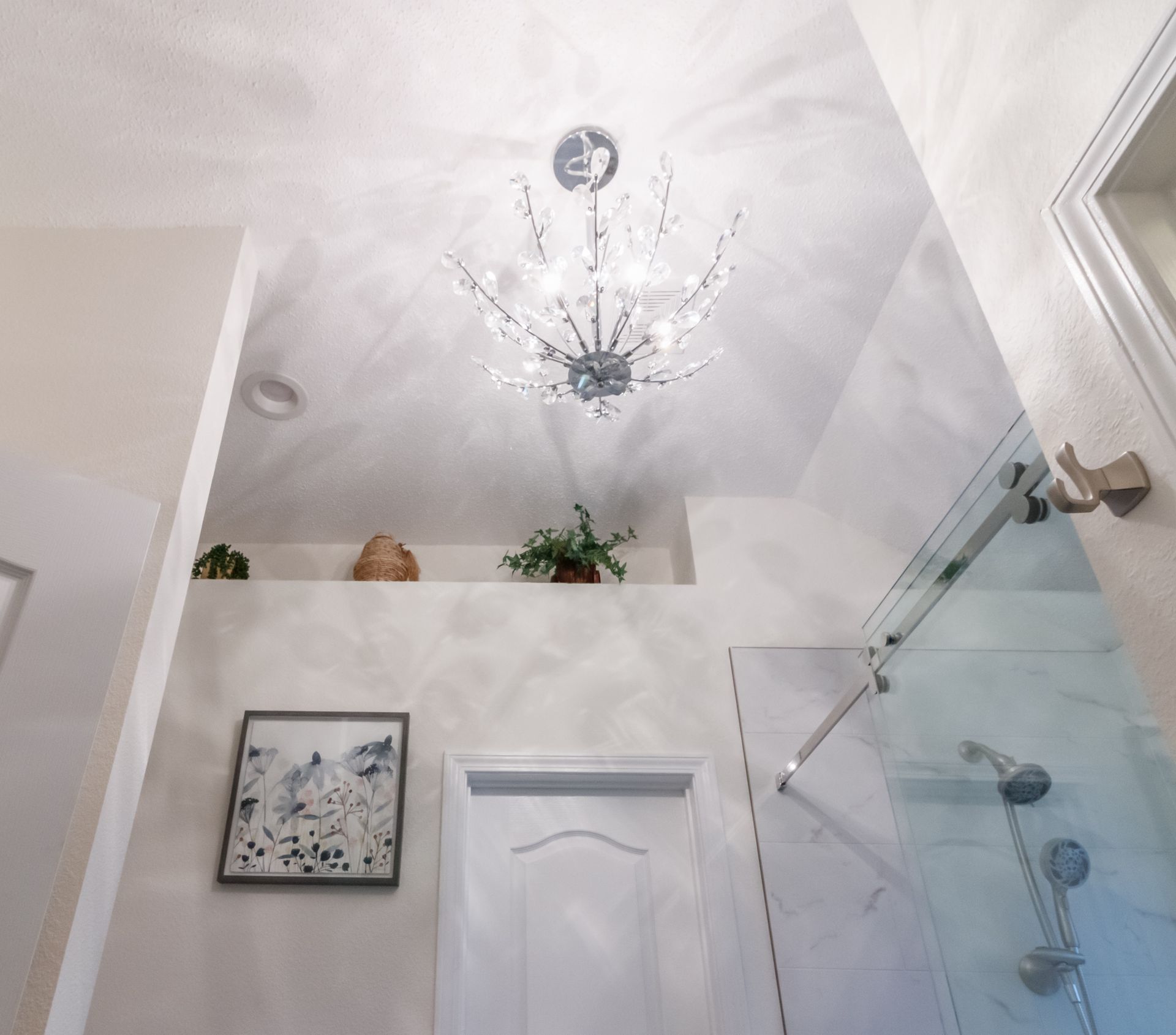 Bright bathroom with a crystal chandelier, white walls, a glass shower, and a framed picture above the door