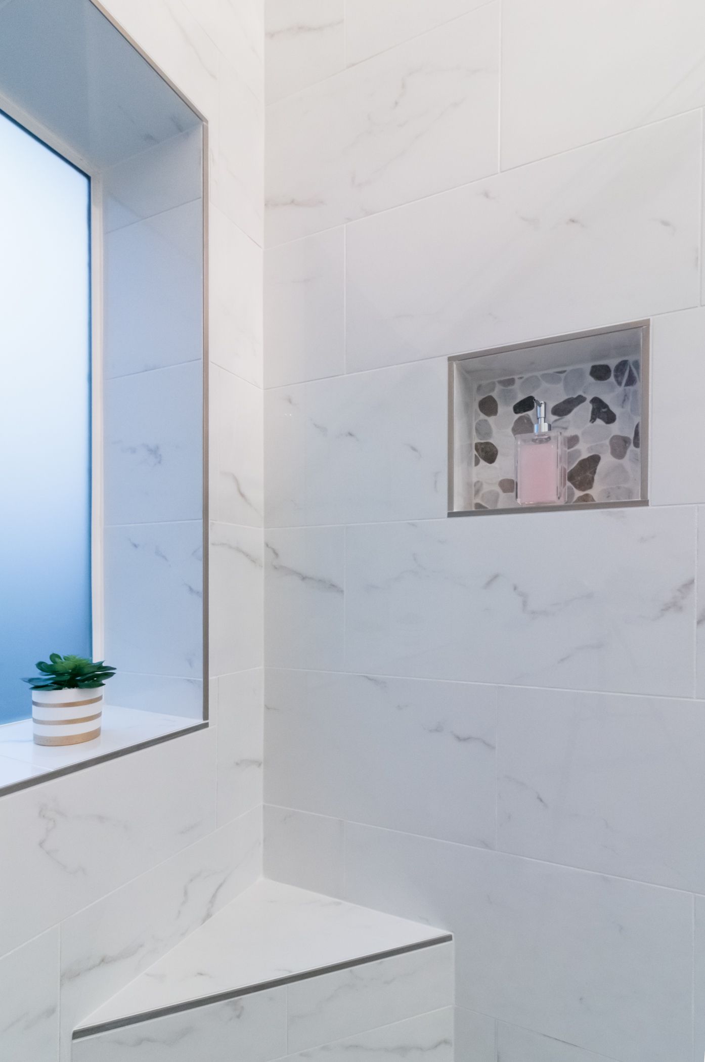 White marble bathroom corner with a window, small potted plant, and recessed wall shelf.
