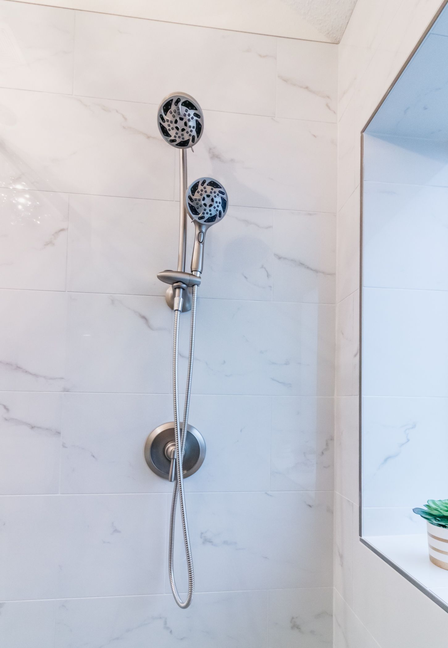 Chrome shower fixture on white tiled wall beside a window niche with a small plant.