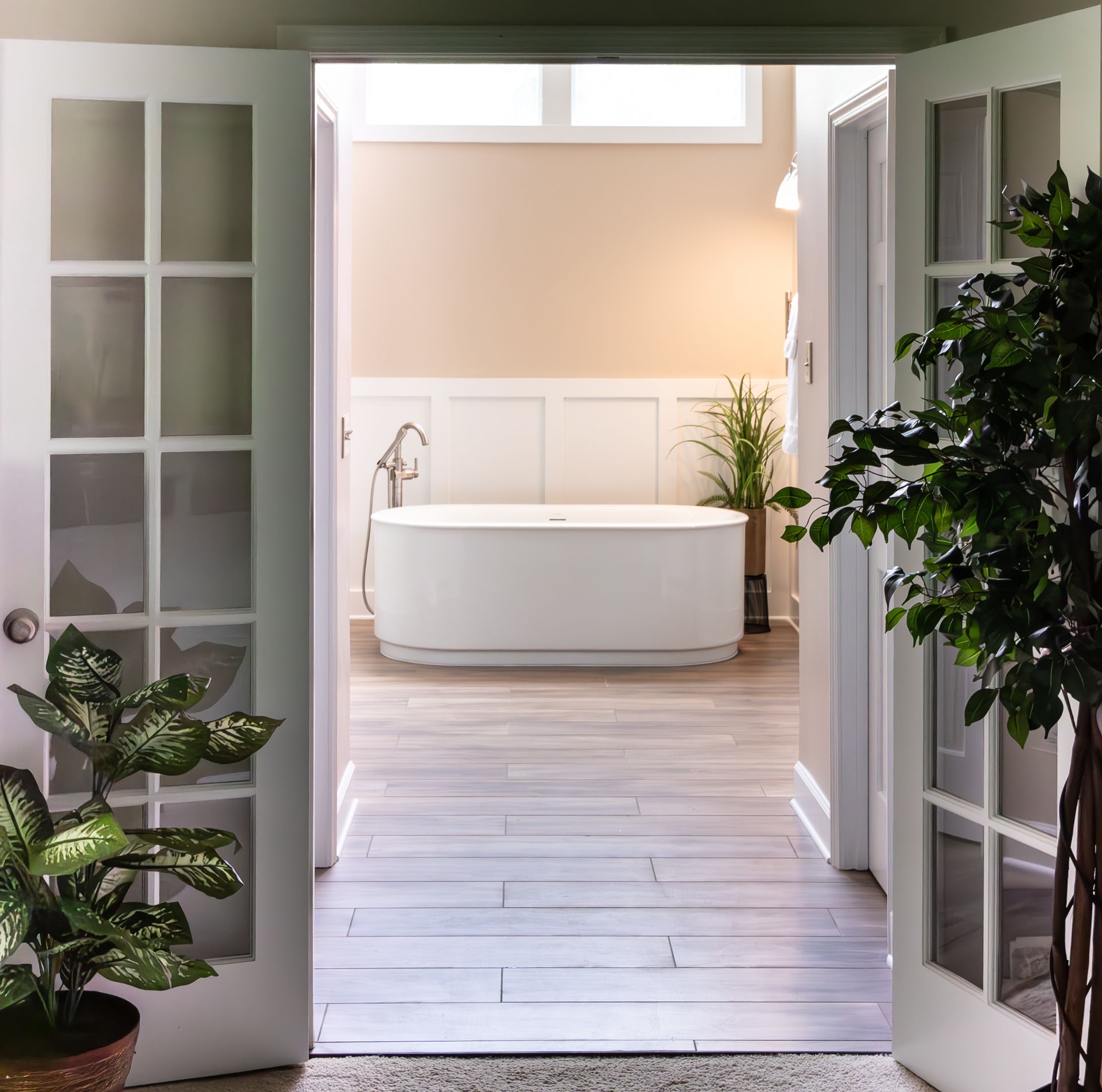 Open French doors reveal a bright, modern bathroom with a freestanding tub and plants