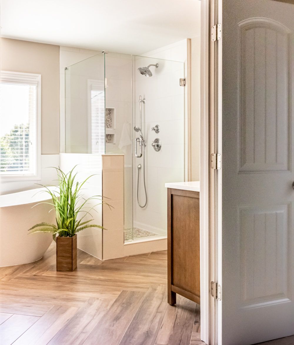 Bright bathroom with walk-in shower, wood vanity, and a potted plant on herringbone wood floor.