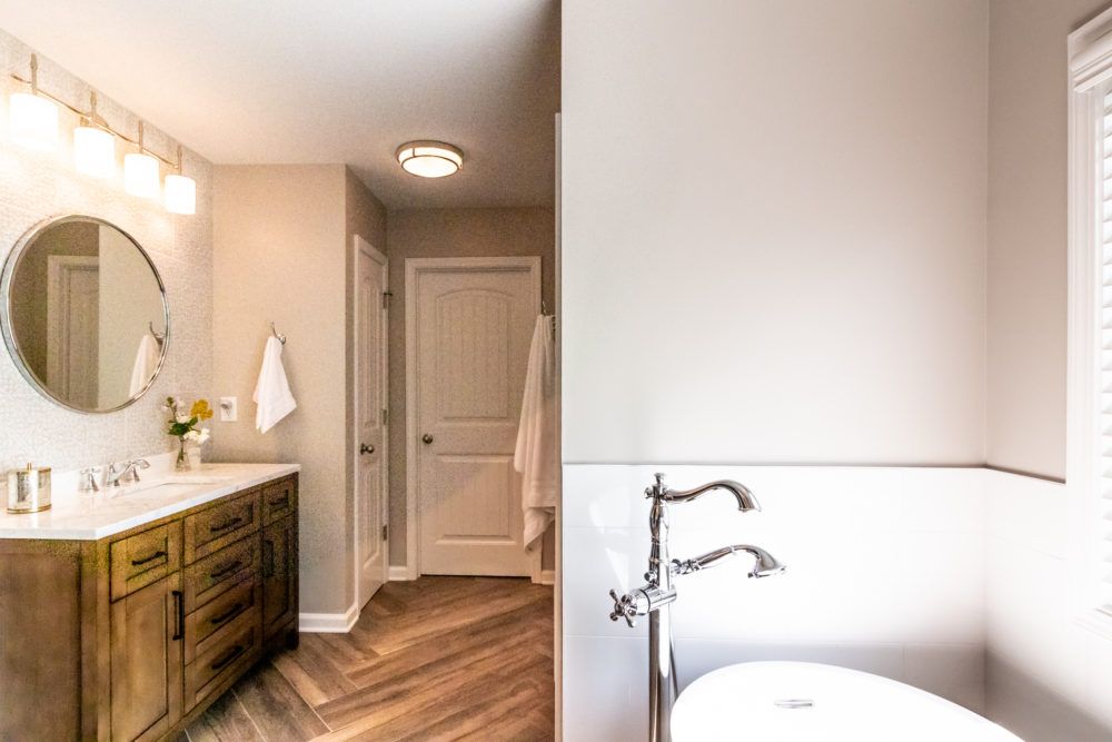 Modern bathroom with wood vanity, round mirror, lit sconces, and a partially visible tub.