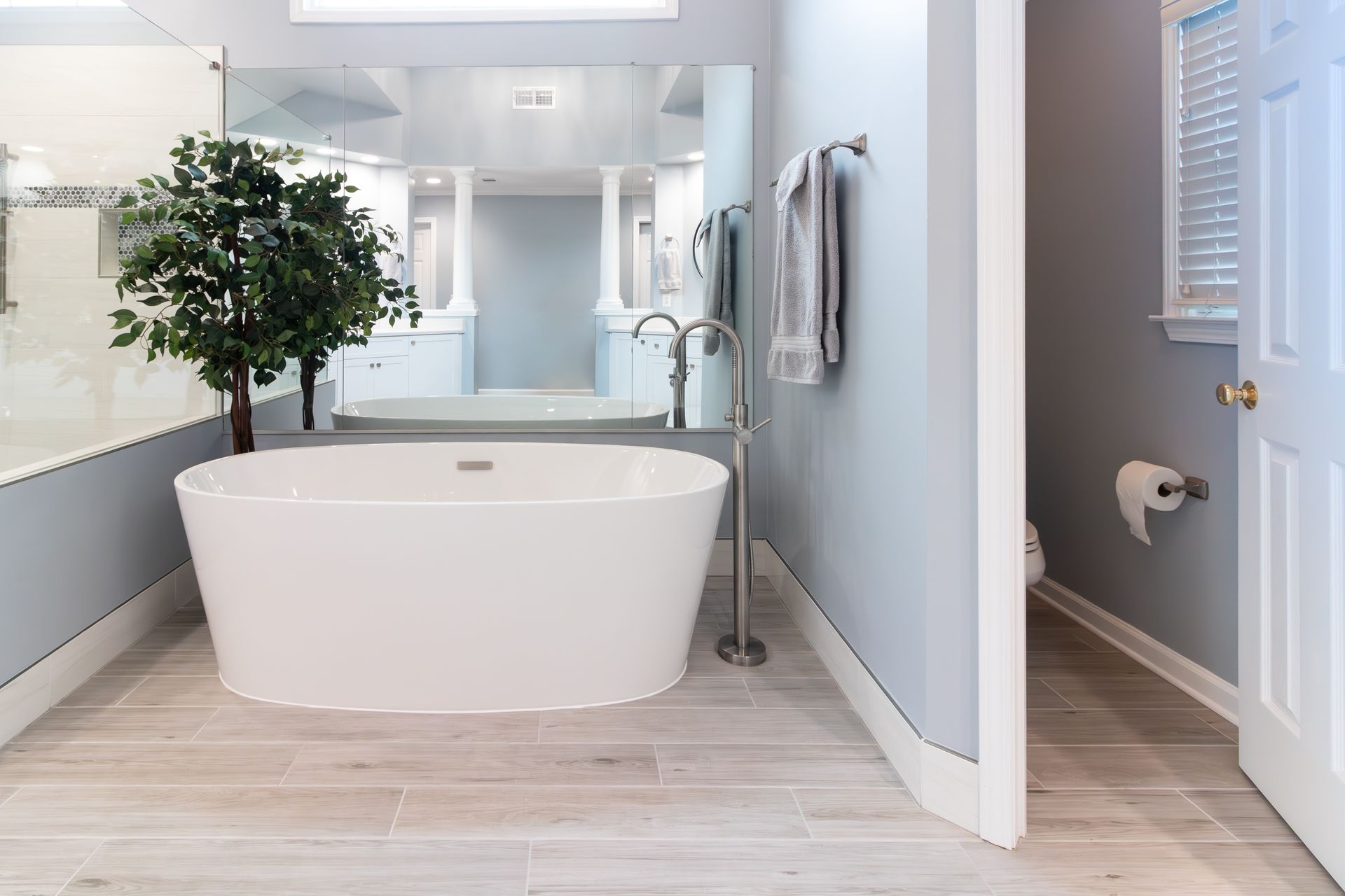 Bright modern bathroom with a freestanding white tub, large mirror, plant, and open door to a toilet area