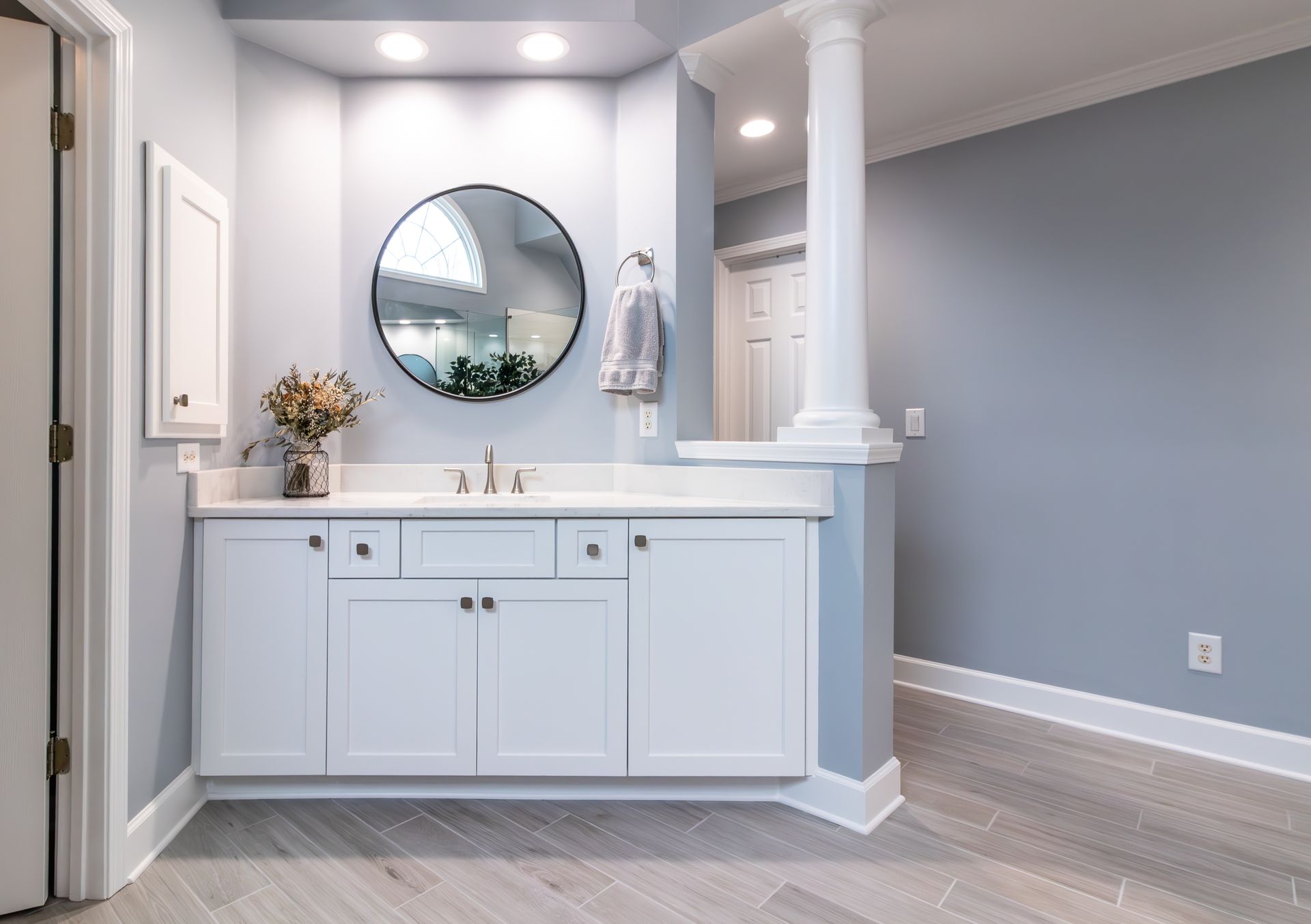 Bright bathroom vanity with round mirror, white cabinets, and light gray walls and flooring