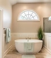 Bright beige bathroom with a white soaking tub under an arched window and a potted plant.