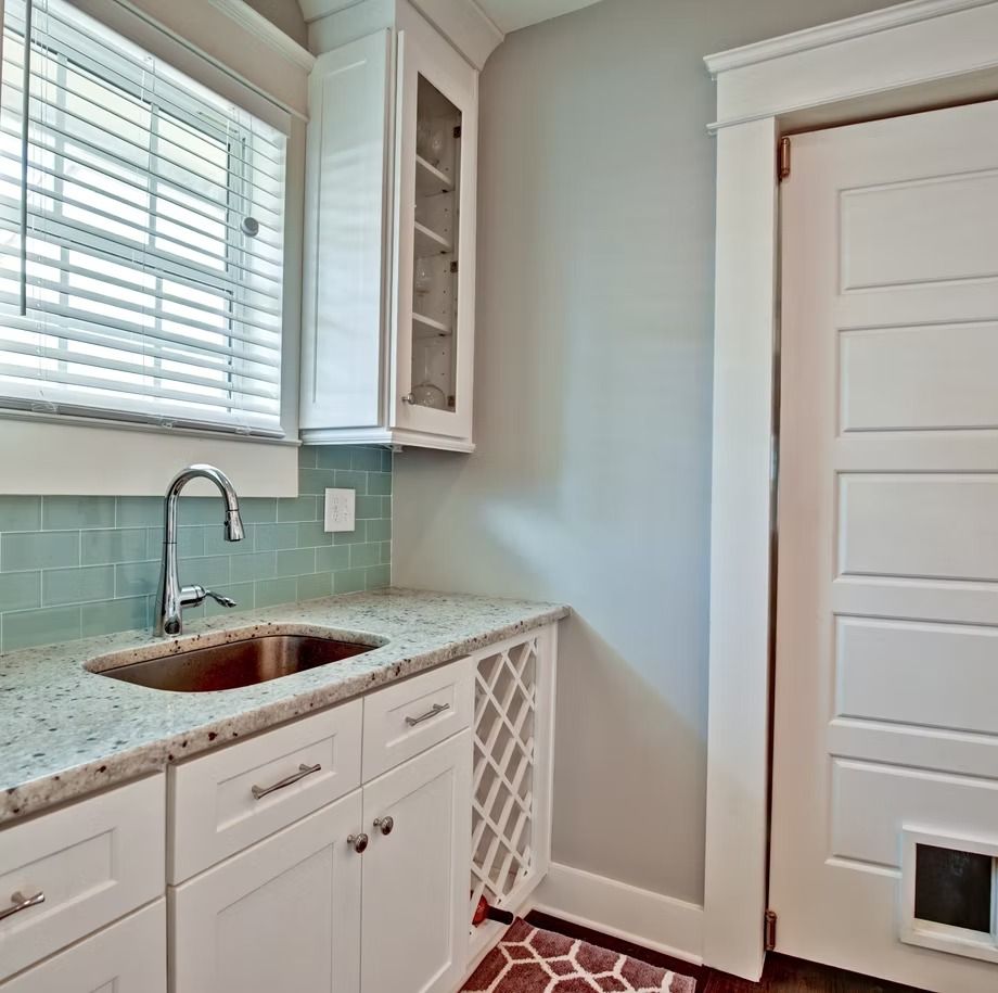 Bright laundry room with white cabinets, granite sink, mint backsplash, and a white door.
