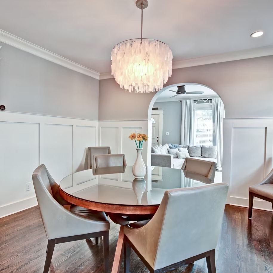 Elegant dining room with round glass table, upholstered chairs, and a chandelier, opening into a bright sitting area