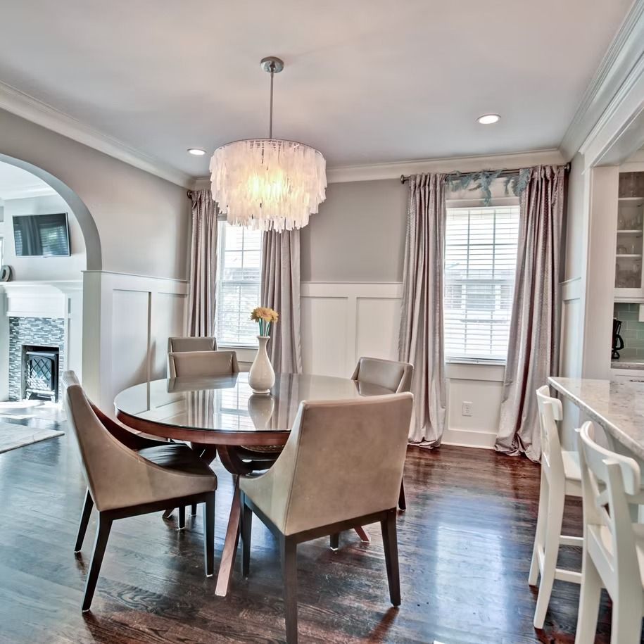Elegant dining room with oval table, upholstered chairs, chandelier, and tall windows with drapes