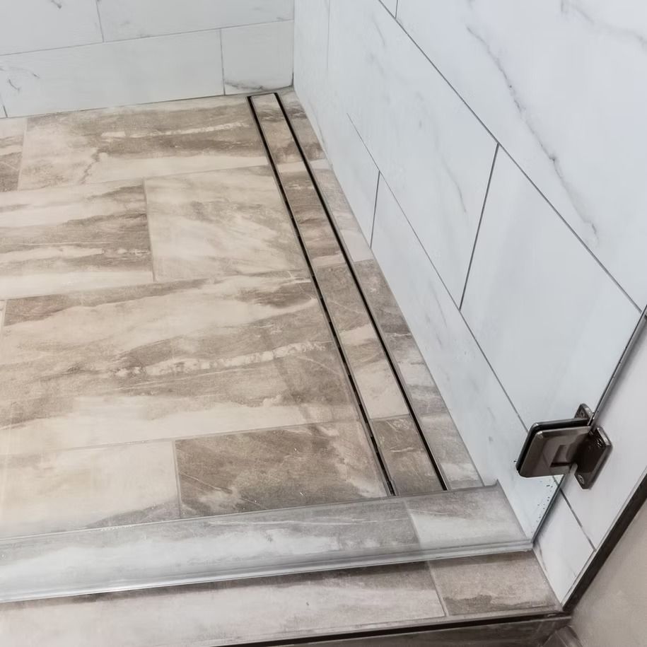 Tiled shower floor with a narrow linear drain and glass door hinge visible.
