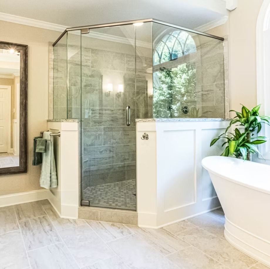 Glass-enclosed tiled shower with marble-look floor, white half wall, and a potted plant in a bright bathroom