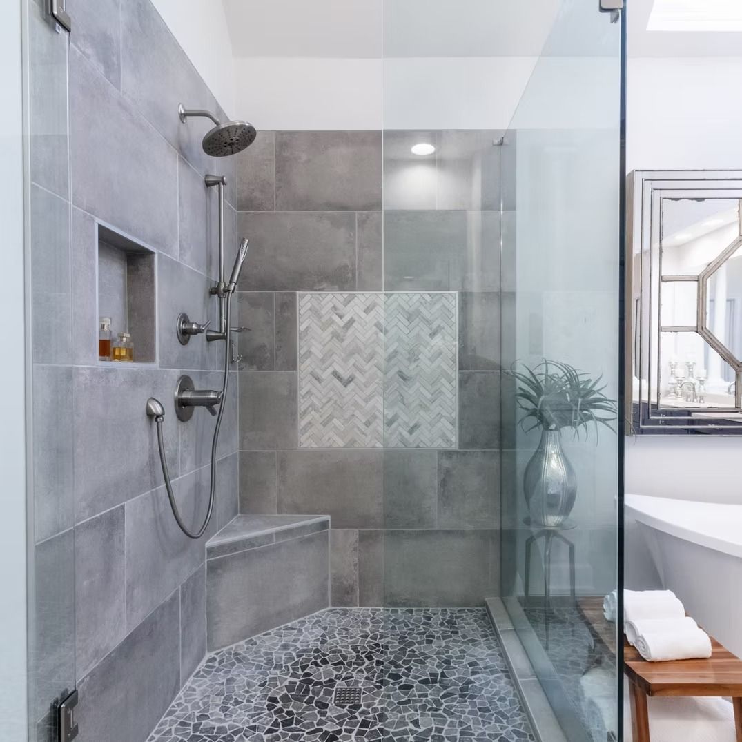 Modern gray tiled bathroom shower with glass partition, rainfall showerhead, and pebble floor.