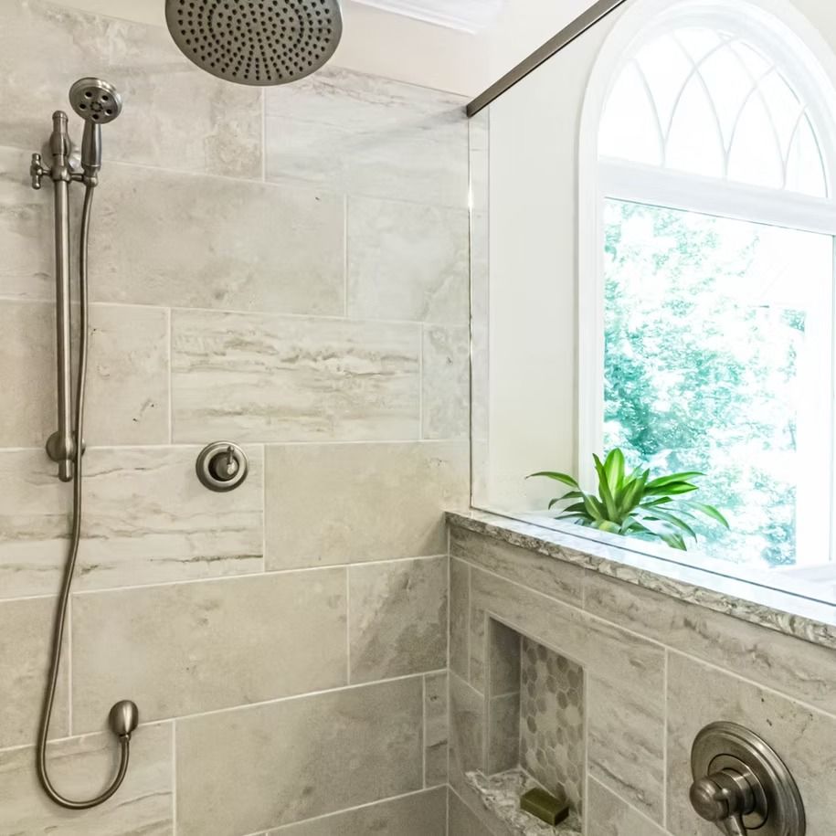 Walk-in shower with stone tile, rain showerhead, built-in shelf, and window ledge with a potted plant.