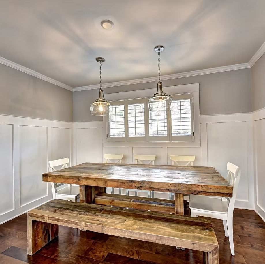 Bright dining room with rustic wooden table and bench, white wainscoting, and two pendant lights