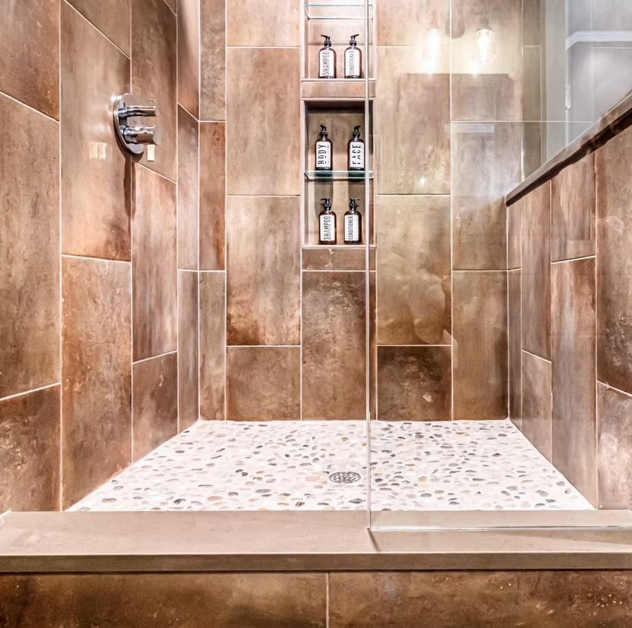 Brown-tiled walk-in shower with a glass partition, built-in niche, and white pebble shower floor.