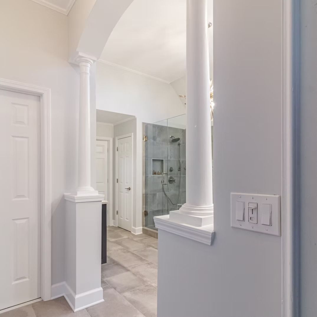 Bright hallway with white columns, arched ceiling, tiled floor, and a glass shower in the background