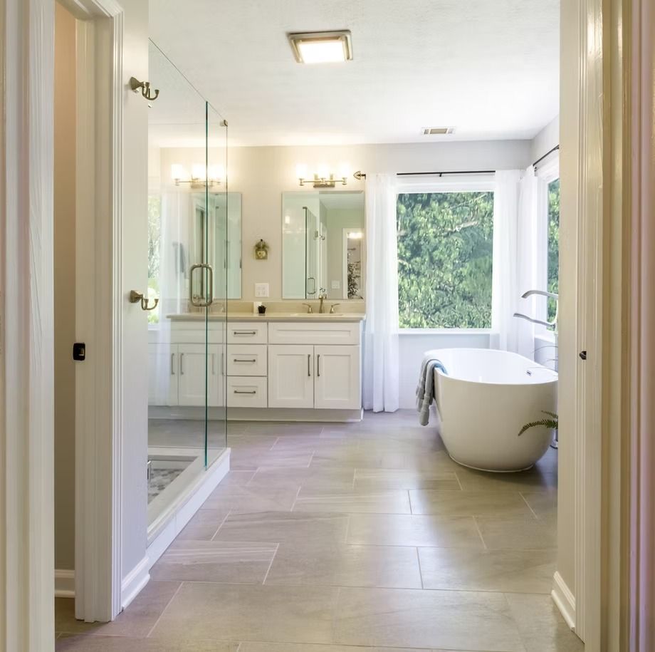 Bright bathroom with glass shower, vanity, freestanding tub, and large window overlooking greenery