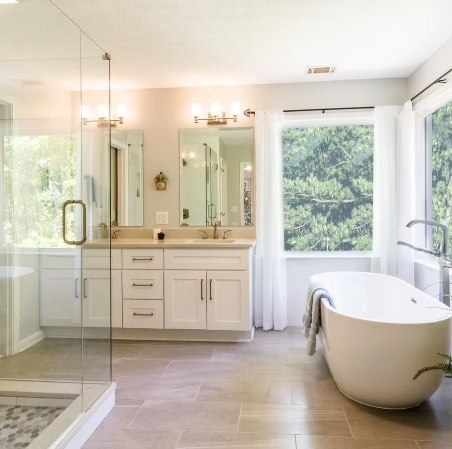 Bright bathroom with double vanity, glass shower, freestanding tub, and large windows overlooking trees