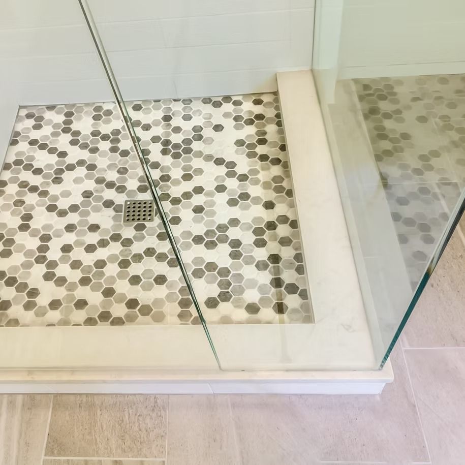 Corner of a glass-enclosed shower with pebble tile floor and a drain on beige tile bathroom floor