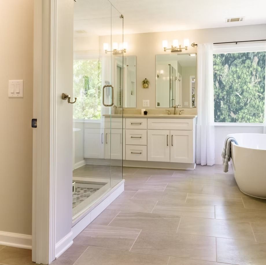 Bright modern bathroom with glass shower, double vanity, large window, and freestanding tub