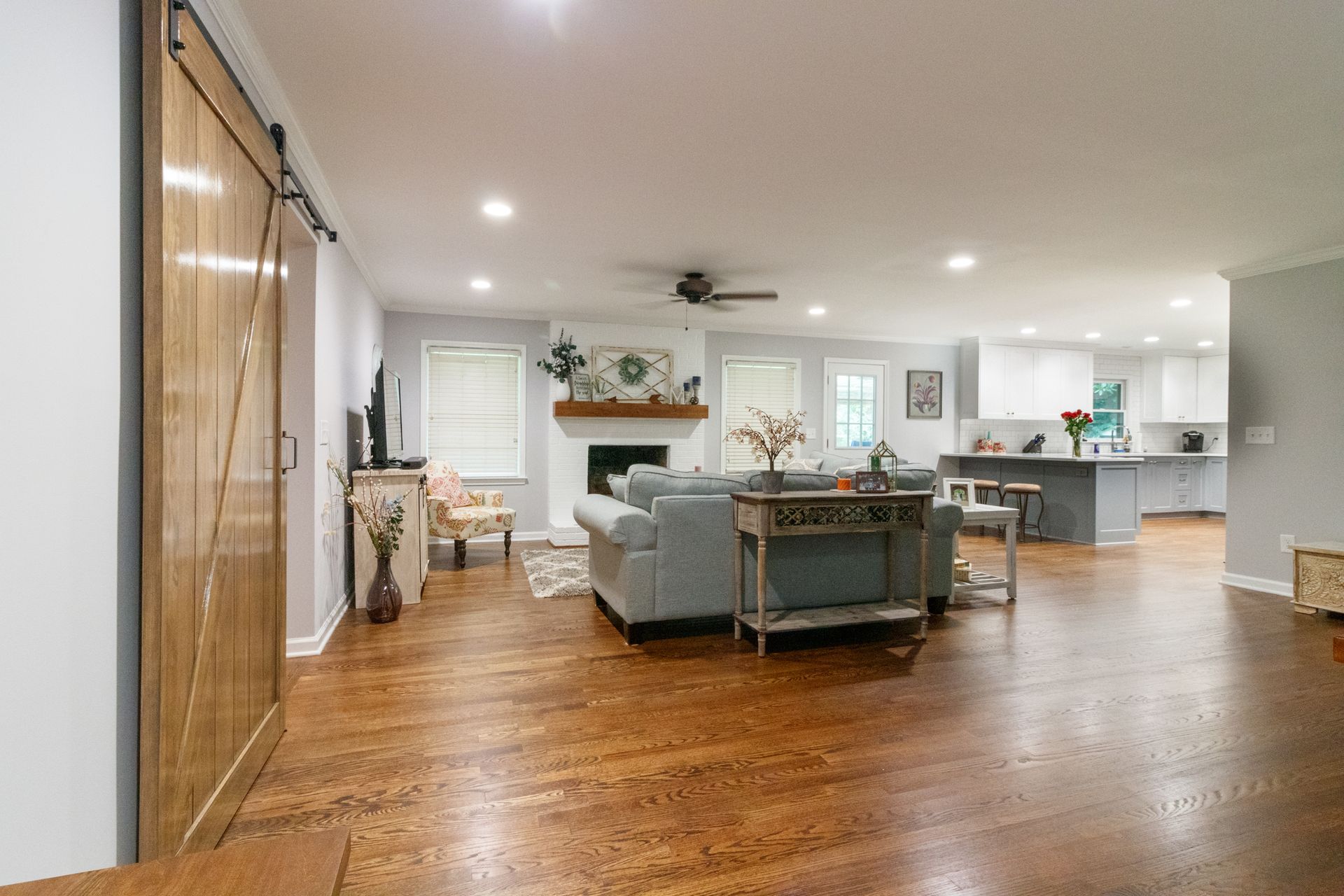 Open-plan living room with wood floors, gray sectional sofa, fireplace, and kitchen in the background
