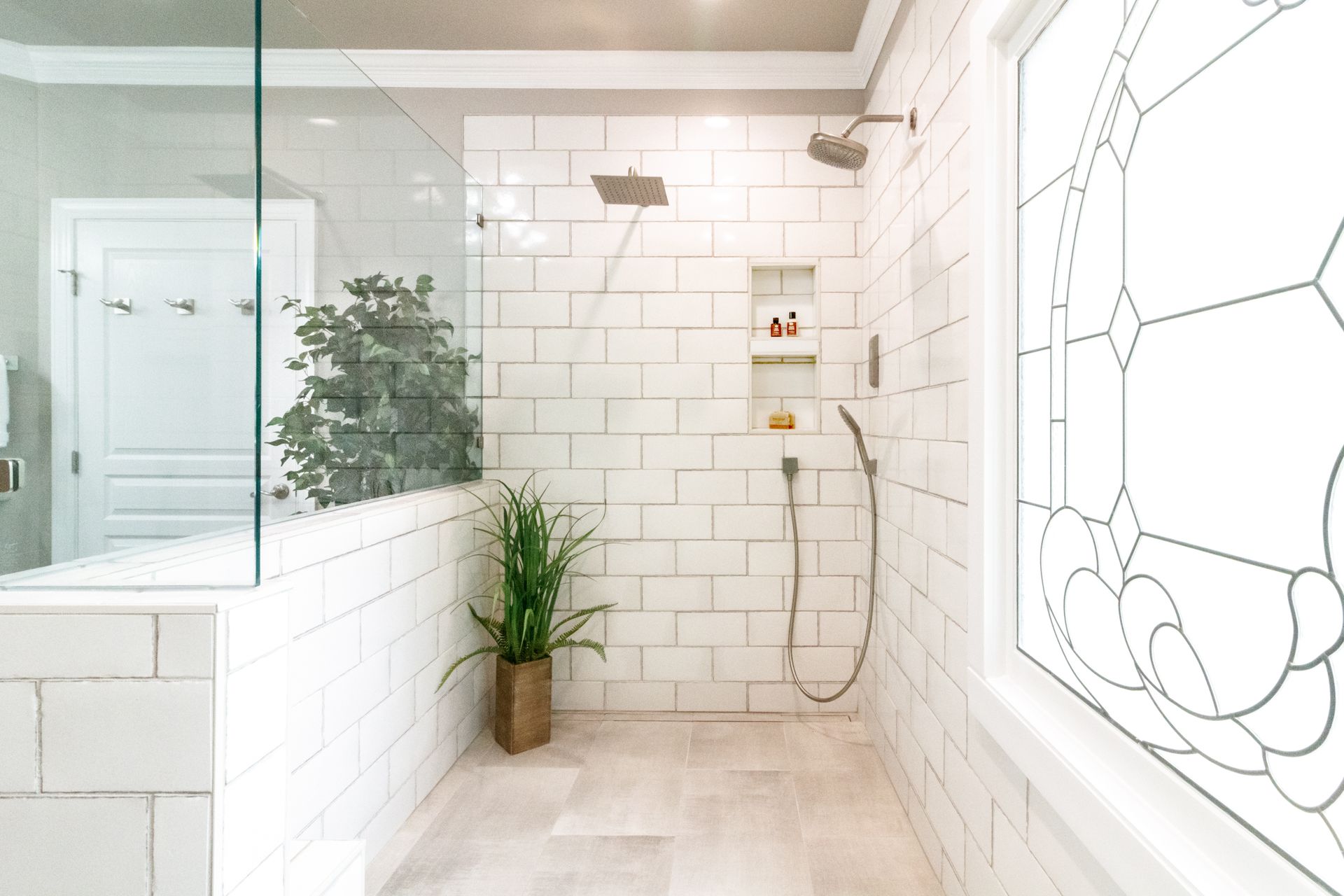 Bright white tiled bathroom with glass shower, potted plant, and decorative window panel
